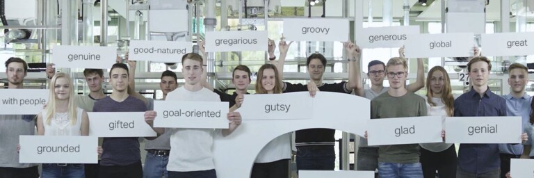 Eine Gruppe junger Menschen steht zusammen und hält Schilder mit positiven Adjektiven, während sie vor einem grossen Buchstaben "G" posieren.