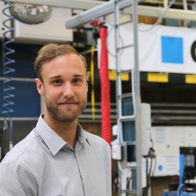 A young man with short hair, wearing a light grey shirt, standing next to machinery in a workshop environment.