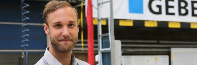 A man in a light gray shirt stands in a workshop, smiling, with industrial equipment and a banner in the background.