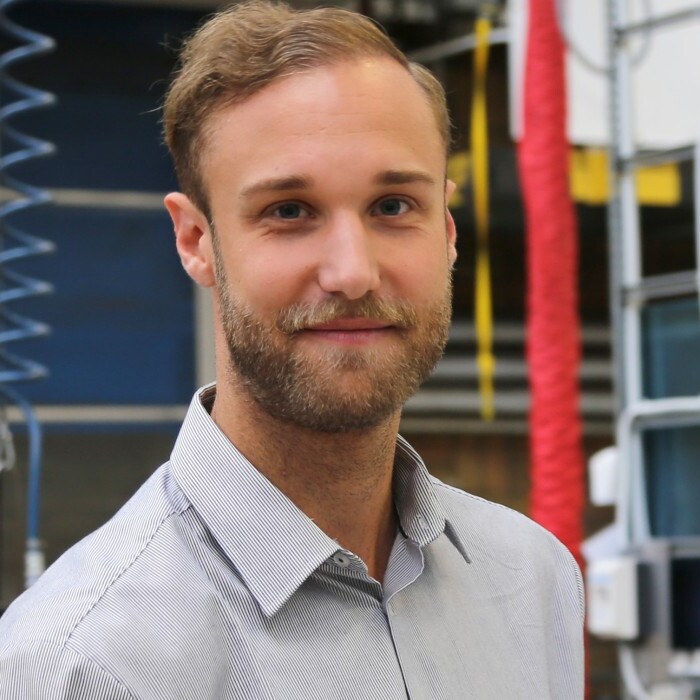 A man in a light gray shirt stands in a workshop, smiling, with industrial equipment and a banner in the background.