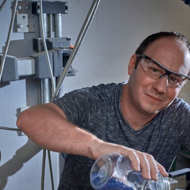 A man wearing safety glasses is pouring a blue liquid from a beaker in a laboratory setting.