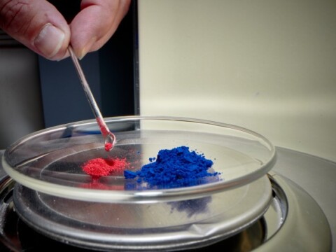 A hand holding a small tool is adding red powder to a dish containing blue and red powders.