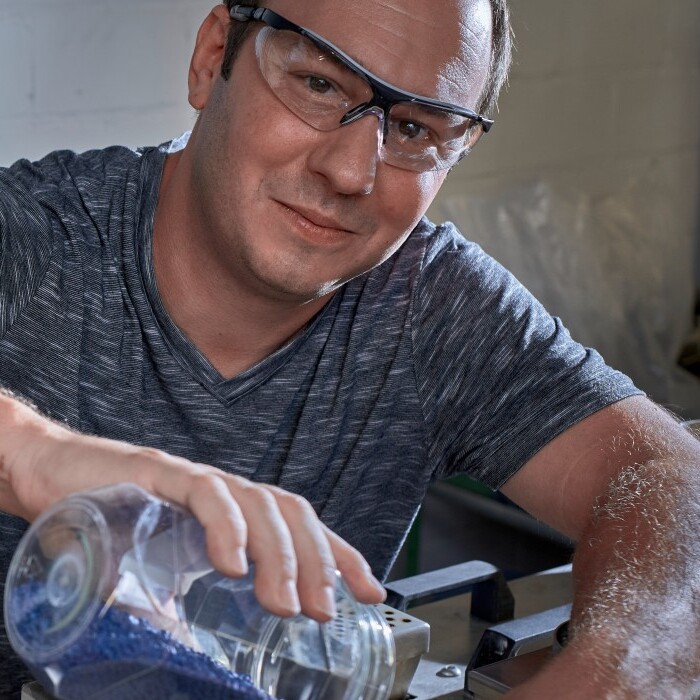 A man wearing safety glasses and a gray shirt pours liquid from a glass bottle into a container in a laboratory setting.
