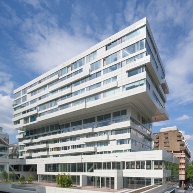 A modern, multi-story building with a unique angular design, large windows, and greenery.