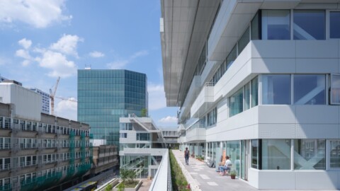 Modern architectural buildings with large glass windows under a clear blue sky, featuring greenery on a balcony.