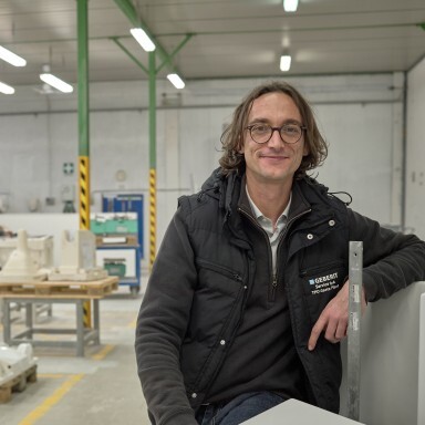 Ein lächelnder Mann mit langen, lockigen Haaren steht in einer Werkstatt und lehnt an einer weissen Sanitärkeramikkonzeption.