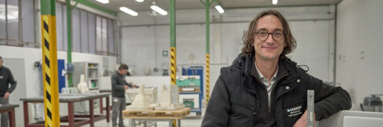 Ein lächelnder Mann mit lockigem Haar steht in einer Fabrikhalle, während im Hintergrund mehrere Personen an Maschinen arbeiten.