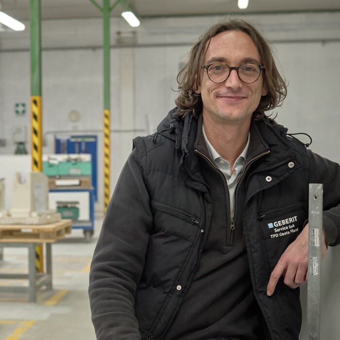 Ein lächelnder Mann mit lockigem Haar steht in einer Fabrikhalle, während im Hintergrund mehrere Personen an Maschinen arbeiten.