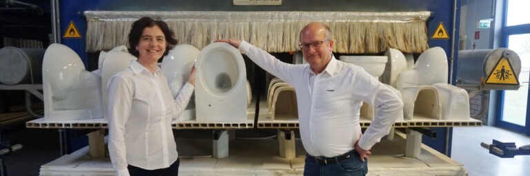 Ein Mann und eine Frau stehen in einer Fabrik, umgeben von WC Keramiken auf einem Produktionsband.
