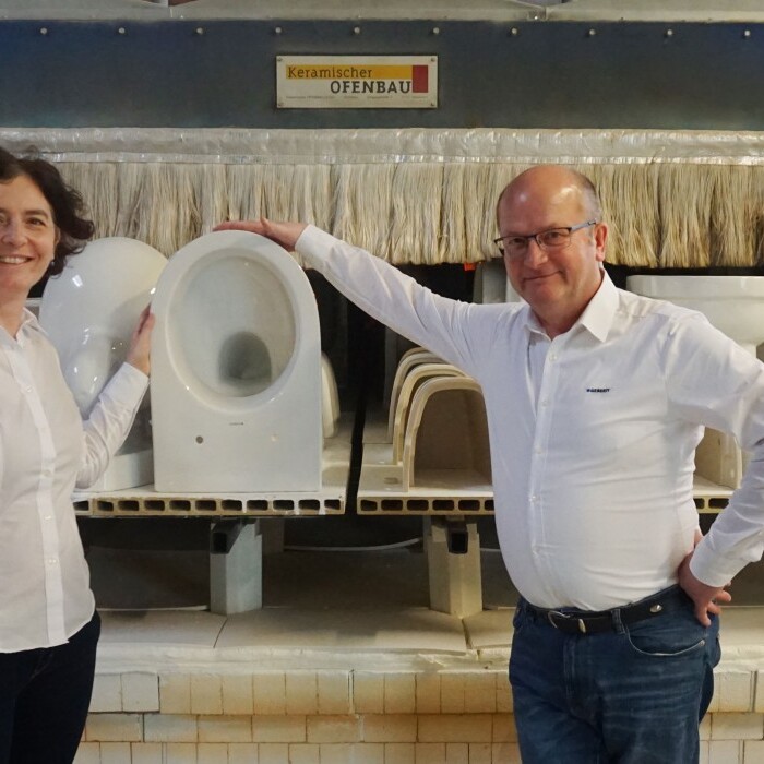 Ein Mann und eine Frau stehen in einer Fabrik, umgeben von WC Keramiken auf einem Produktionsband.