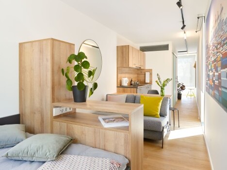 Ein modernes Wohnzimmer mit Holzmöbeln, einer Pflanze, einem runden Spiegel und einem Wohnbereich mit Sofa und Küchenzeile.