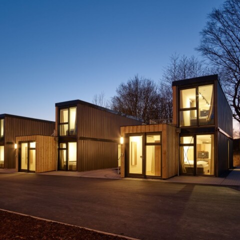 Moderne, rechteckige Holzgebäude mit grossen Fenstern, umgeben von Bäumen, im Abendlicht.