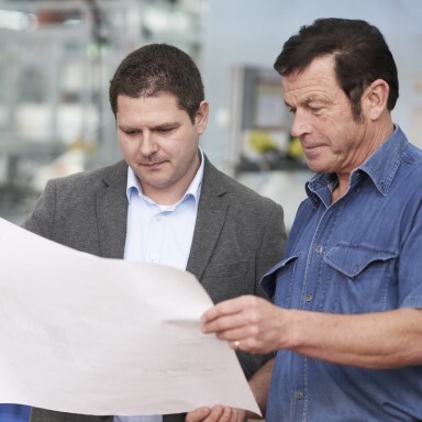 Two men are closely examining a large sheet of paper in an industrial setting, appearing to discuss plans.