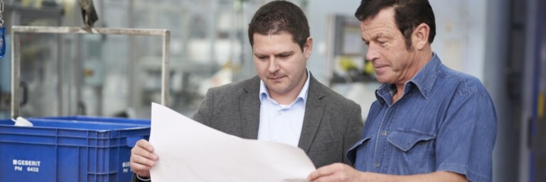 Two men discussing plans over a large sheet of paper in a workshop setting, with crates in the background.