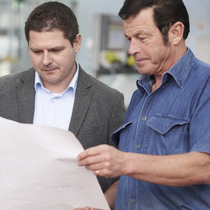 Two men discussing plans over a large sheet of paper in a workshop setting, with crates in the background.