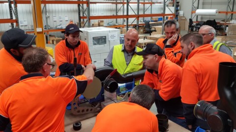 Eine Gruppe von Personen in orangefarbenen T-Shirts, die um einen Tisch stehen und gemeinsam an einem technischen Gerät arbeiten.