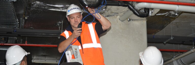 Ein Techniker in orangefarbener Warnweste arbeitet an einer Rohrleitung, während zwei Kollegen in Sicherheitshelmen zuschauen.