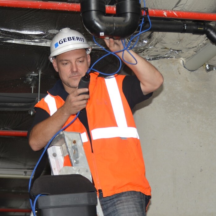 Ein Techniker in orangefarbener Warnweste arbeitet an einer Rohrleitung, während zwei Kollegen in Sicherheitshelmen zuschauen.