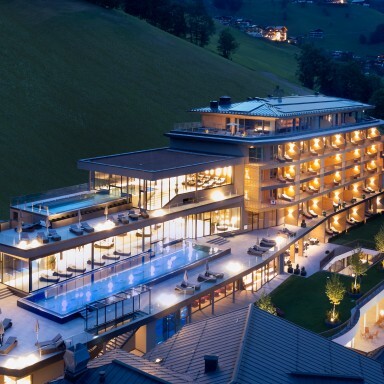 Moderne Hotelanlage mit beleuchteten Terrassen, Schwimmbad und Blick auf bewaldete Hügel bei Abenddämmerung.