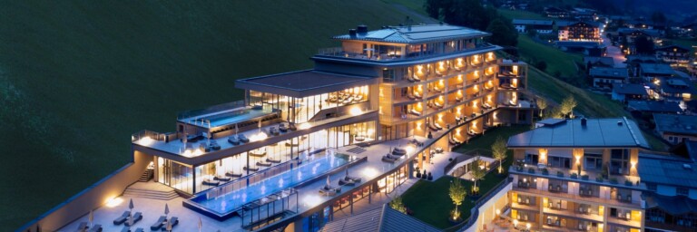 Luxuriöses Hotelgebäude in einer Berglandschaft bei Nacht, mit beleuchteten Balkonen und einem Pool.