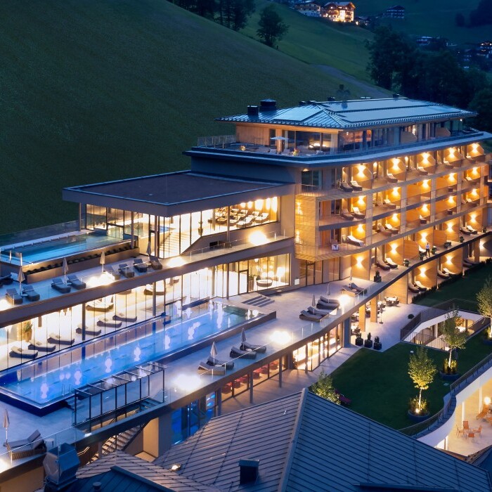 Luxuriöses Hotelgebäude in einer Berglandschaft bei Nacht, mit beleuchteten Balkonen und einem Pool.