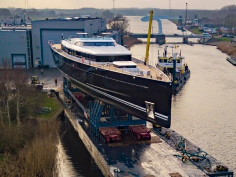 Eine schwarze Yacht auf einem Stützgerüst in einem Hafen, umgeben von Wasser und Werksgebäuden.