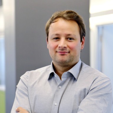 Ein Mann mit kurzem, braunem Haar steht in einem Büro und trägt ein hellblaues Hemd.