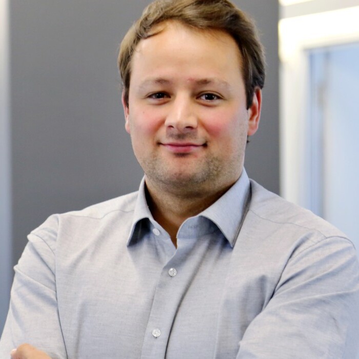 Ein Mann mit kurzem, braunem Haar steht lächelnd in einem Büroraum, gekleidet in einem hellblauen Hemd.