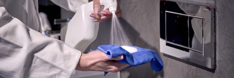 A person in a white lab coat sprays a cleaning solution onto a blue cloth near a wall-mounted actuator plate.