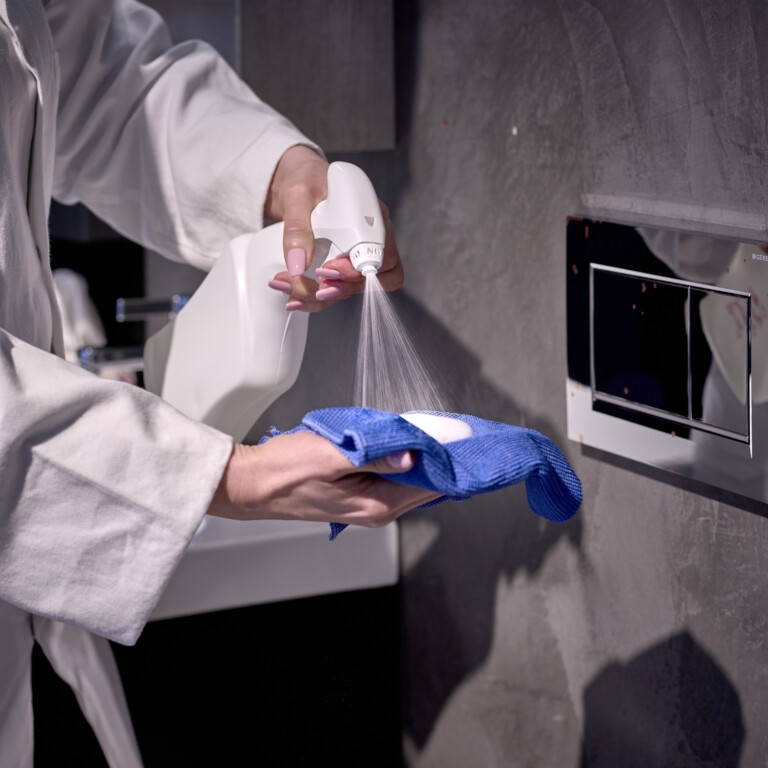 A person in a white lab coat sprays a cleaning solution onto a blue cloth near a wall-mounted actuator plate.