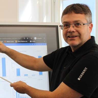 A man with glasses points to a bar graph displayed on a monitor, smiling while standing indoors.