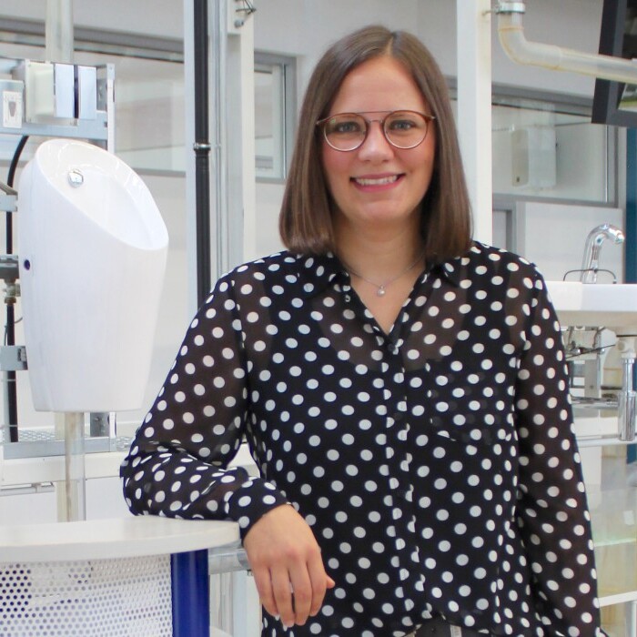 Eine Person mit Brille in einem gepunkteten Hemd steht an einem Tisch, umgeben von sanitären Installationssystemen.