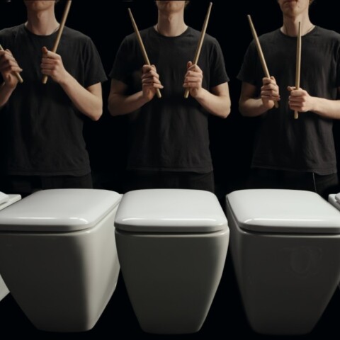 A line of toilets with individuals holding drumsticks poised to play them like drums.