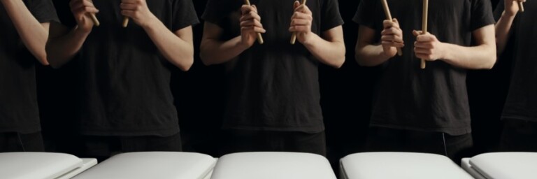 A line of toilets with individuals holding drumsticks poised to play them like drums.