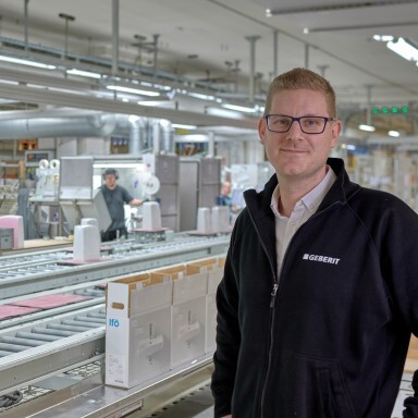 Ein Mann mit Brille und schwarzer Jacke steht im Vordergrund einer Produktionshalle, im Hintergrund sind Maschinen und Mitarbeiter sichtbar.