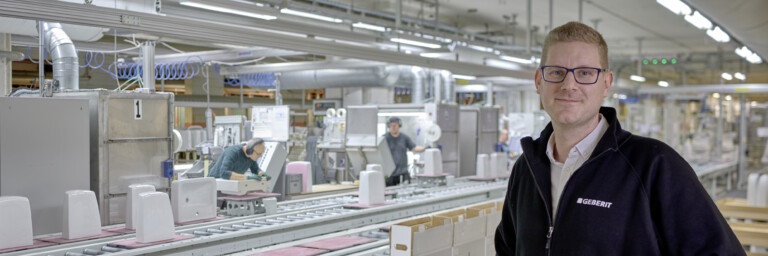 Mann in schwarzer Jacke steht in einer modernen Lager- und Produktionshalle mit Förderband und Mitarbeitern im Hintergrund.