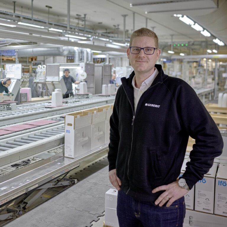 Mann in schwarzer Jacke steht in einer modernen Lager- und Produktionshalle mit Förderband und Mitarbeitern im Hintergrund.