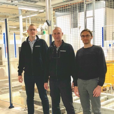 Three men standing in a factory setting with industrial equipment in the background.