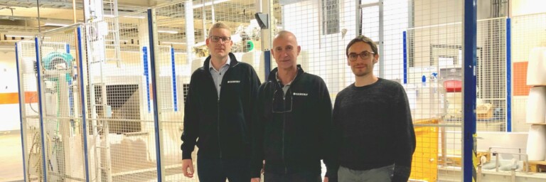 Three men standing in an industrial setting, with machinery and fencing in the background.