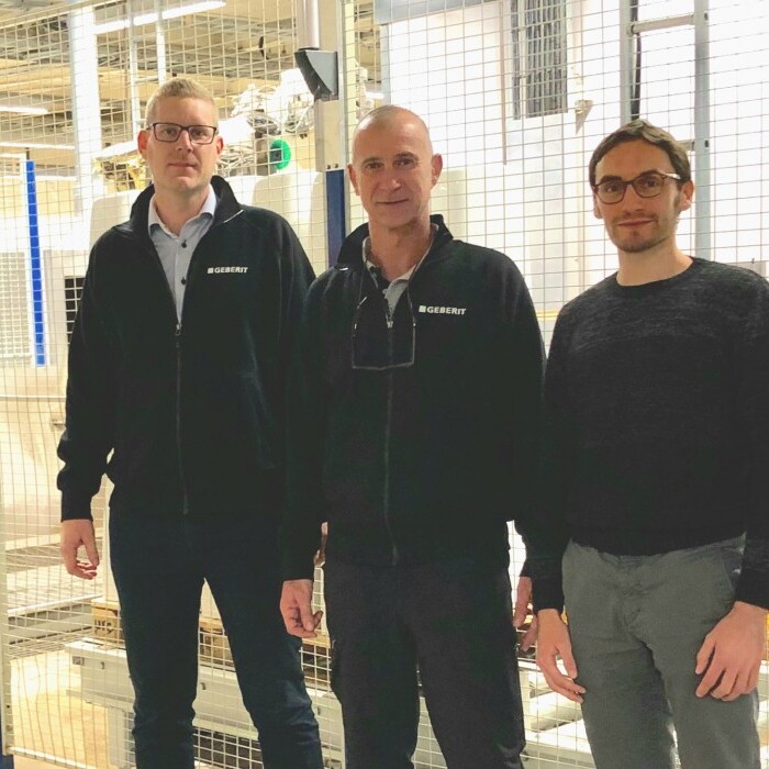 Three men standing in an industrial setting, with machinery and fencing in the background.