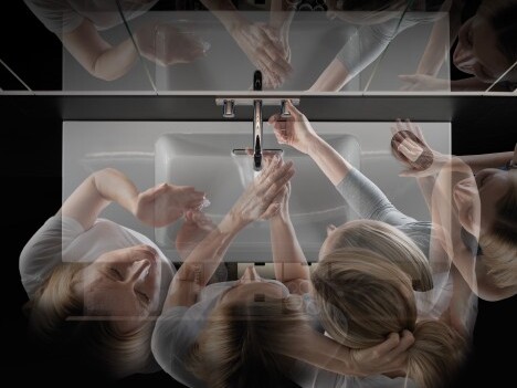A top-down view of multiple overlapping hands reaching towards a sink faucet, creating a sense of movement.