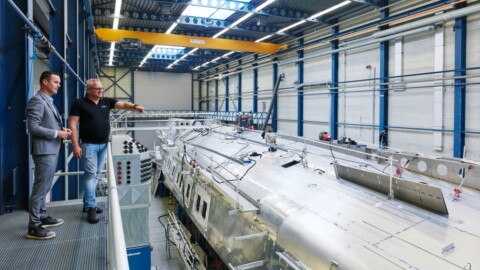 Zwei Personen in einem Industriegebäude stehen auf einem Balkon, während sie auf ein grosses, glänzendes Objekt darunter zeigen.