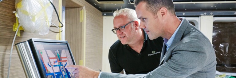 Zwei Männer stehen an einem Bildschirm und betrachten eine technische Ansicht, während einer von ihnen mit dem Finger auf den Bildschirm zeigt.