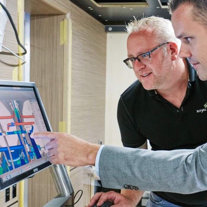 Zwei Männer stehen an einem Bildschirm und betrachten eine technische Ansicht, während einer von ihnen mit dem Finger auf den Bildschirm zeigt.