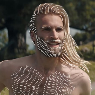 A man with long blonde hair and a unique patterned beard and chest, set against a natural outdoor background.