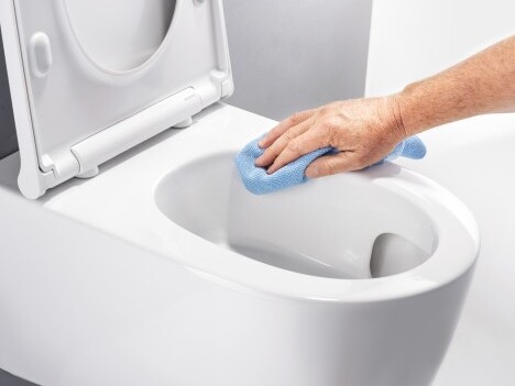 A person's hand using a blue cleaning cloth to wipe the rim of a white toilet bowl.
