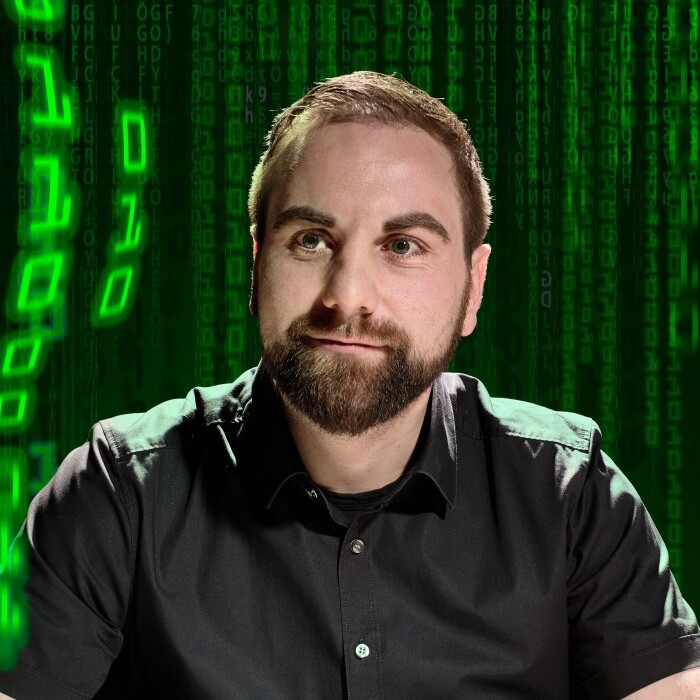 A man with a beard sits in front of a green digital background with zeros and ones.