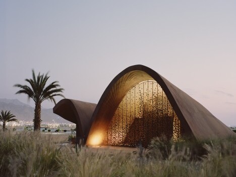 Moderne, geschwungene Architektur mit beleuchtetem Innenraum, umgeben von Palmen und Graslandschaft bei Dämmerung.