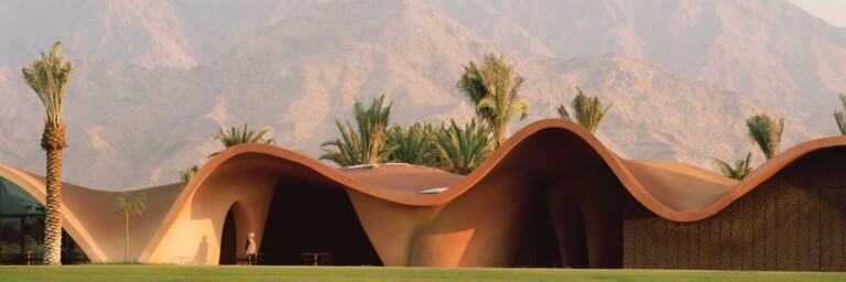 Auf einem Grundstück steht ein modernes, wellenförmiges Gebäude mit einer erdigen Farbgestaltung, umgeben von Palmen, vor einer bergigen Landschaft.