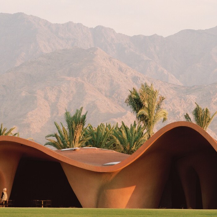 Auf einem Grundstück steht ein modernes, wellenförmiges Gebäude mit einer erdigen Farbgestaltung, umgeben von Palmen, vor einer bergigen Landschaft.